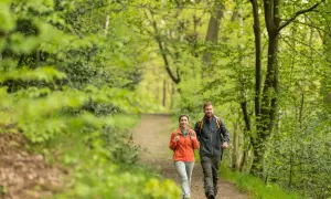 Ein Paar genießt eine Wanderung mit Einkehr im Grünen. Sie gehen auf einem Waldweg, umgeben von üppiger Vegetation.
