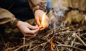 Hände entzünden mit einem Streichholz ein kleines Feuer aus Ästen. Das Lagerfeuer machen beginnt.