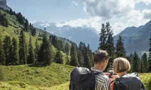 Ein Paar plant seine **Wanderung** in den Alpen. Sie schauen auf ihr Handy, um die Route zu prüfen, umgeben von grünen Hängen und schneebedeckten Gipfeln.
