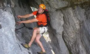 Mann klettert gesichert im toten Gebirge. Er trägt Helm, orangefarbenes Shirt und Rucksack.