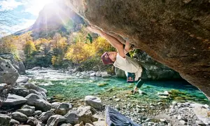 Ein Kletterer meistert eine anspruchsvolle Route beim **Tessin Bouldern** über einem kristallklaren Fluss. Herbstlaub und Berge im Hintergrund.