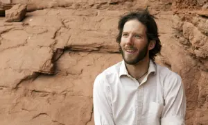 Aron Ralston heute, mit Bart und zerzaustem Haar, blickt lächelnd in die Ferne vor einer roten Felswand.