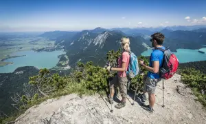 Ein Paar genießt die Aussicht beim Wandern in Mittenwald. Türkisfarbene Seen und grüne Berge erstrecken sich unter ihnen.