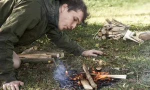 Mann entfacht mit einem Feuerstahl ein Lagerfeuer. Er pustet vorsichtig auf die Flammen, um sie zu nähren.