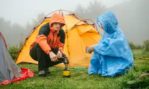 Zwei Personen im Regen bauen ein Zelt auf und bereiten sich ein warmes Getränk zu.