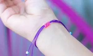 Eine Hand trägt ein lila Armband mit einem doppelten Schiebeknoten. Kleine blaue Perlen zieren die Enden.