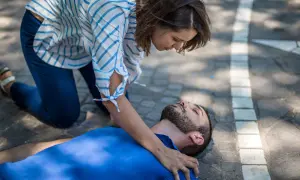 Eine Frau kniet neben einem Mann, der auf dem Boden liegt. Sie prüft seine Atmung, um zu sehen, was passiert bei einem allergischen Schock.