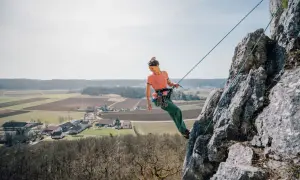 Frau klettert an einem Felsen, Seil ist sicher zum toprope einrichten befestigt. Im Hintergrund Felder und ein Dorf.