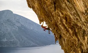 Kletterin hängt an Felswand, die Schwierigkeit der Route wird auf der UIAA-Skala bewertet. Meer und Berge im Hintergrund.