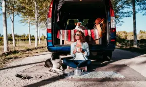 Frau mit Hunden genießt Kaffee vor ihrem Van. Tolle Camping-Hacks für entspannte Ausflüge mit Haustieren.