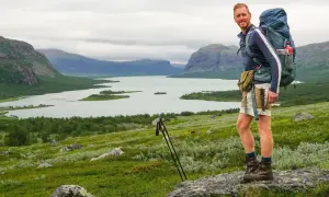 Wanderer mit Rucksack genießt die weite Landschaft im Nationalpark Sarek. Berge, See und grüne Hänge prägen das Bild.