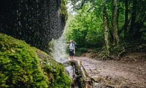 Wanderin unter einem Wasserfall im Schwarzwald. Moosbedeckte Felsen und üppiger Wald säumen den Weg.