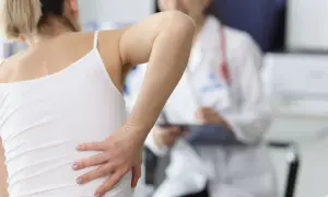 Frau hält sich den Rücken, wo sie eine geprellte Rippe vermutet. Ein Arzt im Hintergrund macht sich Notizen.