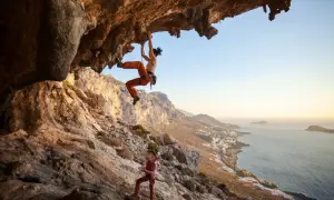 Zwei Frauen beim Klettern an einer Felswand über dem Meer. Die obere Kletterin hängt unter einem Überhang, die untere sichert sie. Verschiedene Kletterarten sind hier zu sehen.
