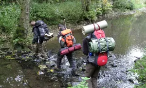 Drei Wanderer durchqueren einen Bach. Ein Mann befestigt seinen Schlafsack am Rucksack.