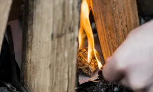 Holzscheite umgeben ein kleines Bündel Zunder, das gerade entzündet wird. Die Flammen tanzen und beginnen, das Holz zu verzehren. Ein Finger ist im Vordergrund zu sehen, der das Feuer macht.