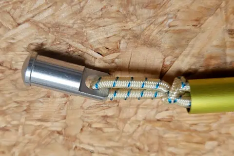 Detailaufnahme: Ein silbernes Endstück mit einer Öse ist mit einem elastischen Seil verbunden, das in ein gelbes Rohr führt. Dies ist eine Methode, um eine Zeltstange zu reparieren.