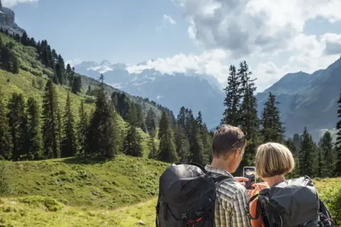 Ein Paar plant seine **Wanderung** in den Alpen. Sie schauen auf ihr Handy, um die Route zu prüfen, umgeben von grünen Hängen und schneebedeckten Gipfeln.