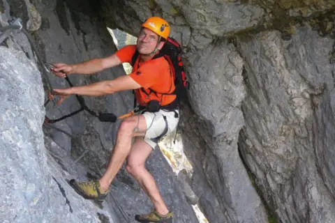 Mann klettert gesichert im toten Gebirge. Er trägt Helm, orangefarbenes Shirt und Rucksack.
