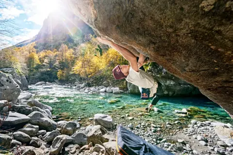 Ein Kletterer meistert eine anspruchsvolle Route beim **Tessin Bouldern** über einem kristallklaren Fluss. Herbstlaub und Berge im Hintergrund.