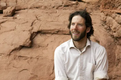 Aron Ralston heute, mit Bart und zerzaustem Haar, blickt lächelnd in die Ferne vor einer roten Felswand.