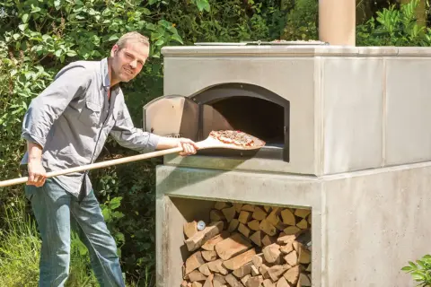 Mann schiebt Pizza in selbstgebauten Outdoor-Ofen. Holz stapelt sich darunter.