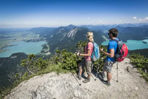 Ein Paar genießt die Aussicht beim Wandern in Mittenwald. Türkisfarbene Seen und grüne Berge erstrecken sich unter ihnen.