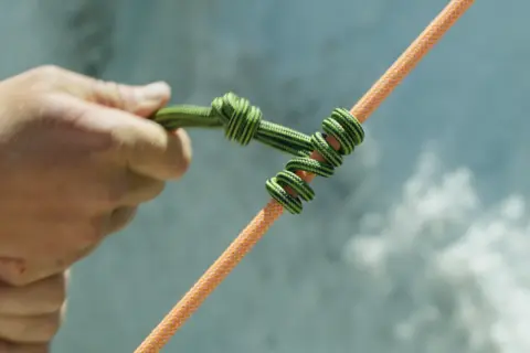 Eine Hand bindet einen HMS Knoten auf ein oranges Seil.