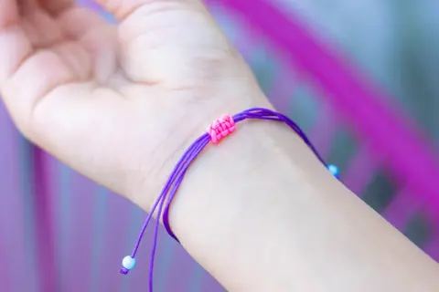 Eine Hand trägt ein lila Armband mit einem doppelten Schiebeknoten. Kleine blaue Perlen zieren die Enden.