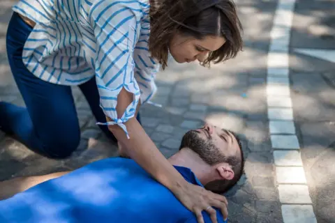 Eine Frau kniet neben einem Mann, der auf dem Boden liegt. Sie prüft seine Atmung, um zu sehen, was passiert bei einem allergischen Schock.