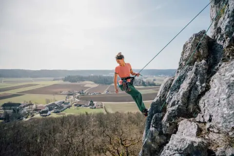 Frau klettert an einem Felsen, Seil ist sicher zum toprope einrichten befestigt. Im Hintergrund Felder und ein Dorf.