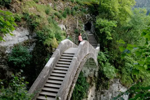 Eine Frau genießt die Aussicht auf einer Steinbrücke, während sie in Inzigkofen wandert. Treppen führen zu einer Höhle.