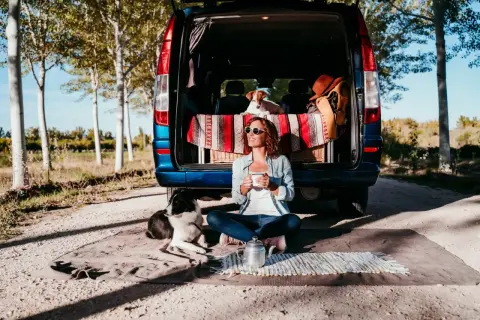 Frau mit Hunden genießt Kaffee vor ihrem Van. Tolle Camping-Hacks für entspannte Ausflüge mit Haustieren.
