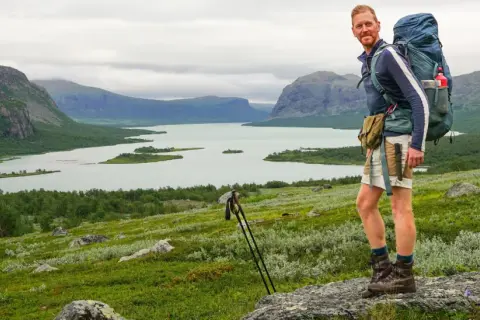 Wanderer mit Rucksack genießt die weite Landschaft im Nationalpark Sarek. Berge, See und grüne Hänge prägen das Bild.