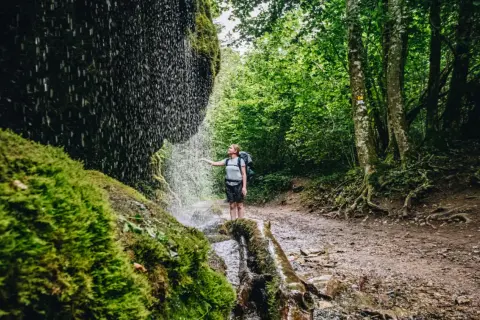 Wanderin unter einem Wasserfall im Schwarzwald. Moosbedeckte Felsen und üppiger Wald säumen den Weg.
