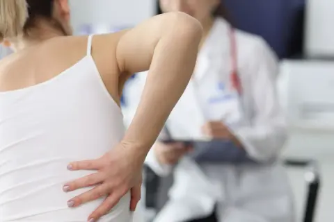 Frau hält sich den Rücken, wo sie eine geprellte Rippe vermutet. Ein Arzt im Hintergrund macht sich Notizen.