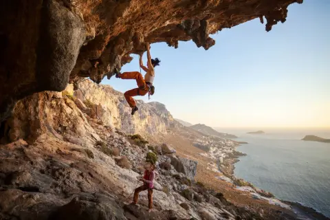 Zwei Frauen beim Klettern an einer Felswand über dem Meer. Die obere Kletterin hängt unter einem Überhang, die untere sichert sie. Verschiedene Kletterarten sind hier zu sehen.