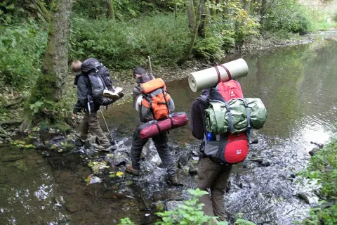 Drei Wanderer durchqueren einen Bach. Ein Mann befestigt seinen Schlafsack am Rucksack.