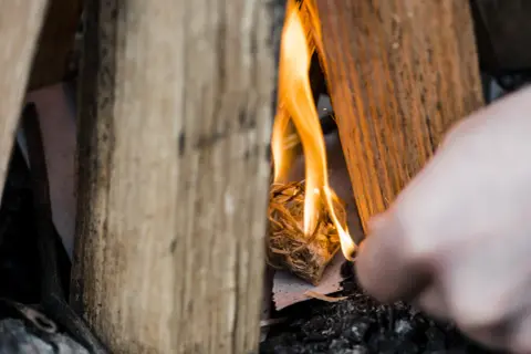 Holzscheite umgeben ein kleines Bündel Zunder, das gerade entzündet wird. Die Flammen tanzen und beginnen, das Holz zu verzehren. Ein Finger ist im Vordergrund zu sehen, der das Feuer macht.