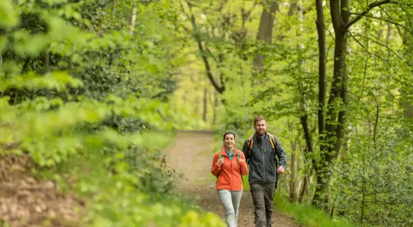 Ein Paar genießt eine Wanderung mit Einkehr im Grünen. Sie gehen auf einem Waldweg, umgeben von üppiger Vegetation.