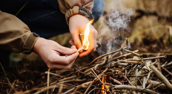 Hände entzünden mit einem Streichholz ein kleines Feuer aus Ästen. Das Lagerfeuer machen beginnt.