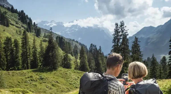 Ein Paar plant seine **Wanderung** in den Alpen. Sie schauen auf ihr Handy, um die Route zu prüfen, umgeben von grünen Hängen und schneebedeckten Gipfeln.