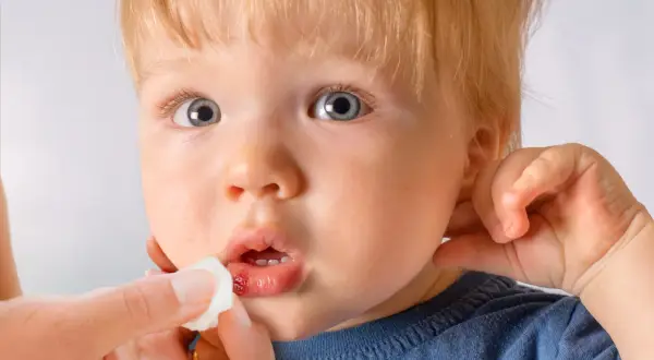 Ein Kind mit blauen Augen und blonden Haaren bekommt die Lippe mit einem Tupfer versorgt. Die Lippe zeigt eine kleine, blutende Platzwunde.
