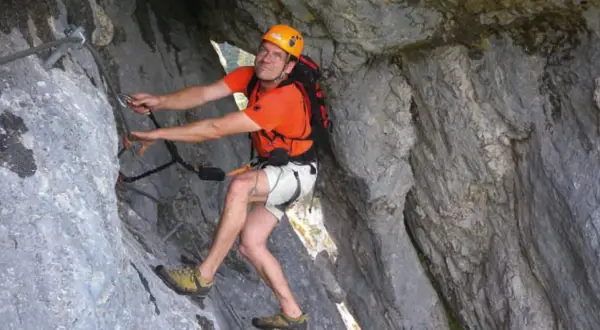 Mann klettert gesichert im toten Gebirge. Er trägt Helm, orangefarbenes Shirt und Rucksack.