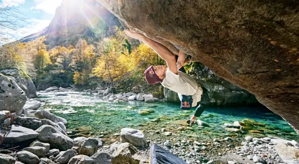 Ein Kletterer meistert eine anspruchsvolle Route beim **Tessin Bouldern** über einem kristallklaren Fluss. Herbstlaub und Berge im Hintergrund.