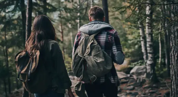 Zwei Personen mit Rucksäcken wandern durch einen Wald. Ein Mann und eine Frau, die sich auf ein Abenteuer vorbereiten, das über einfaches Hiking hinausgeht und eher dem Trekking ähnelt.