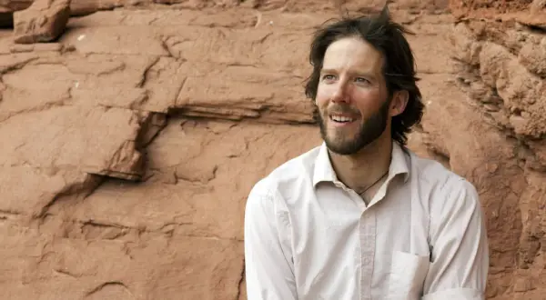 Aron Ralston heute, mit Bart und zerzaustem Haar, blickt lächelnd in die Ferne vor einer roten Felswand.