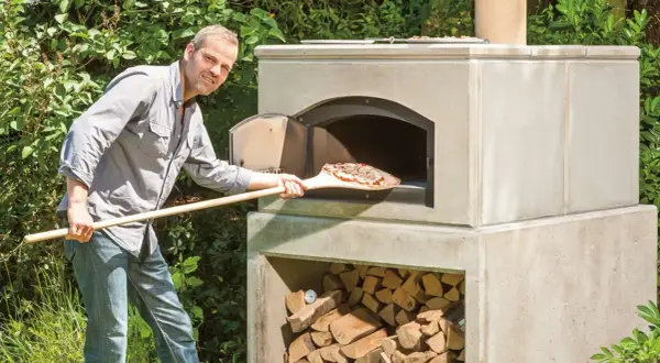 Mann schiebt Pizza in selbstgebauten Outdoor-Ofen. Holz stapelt sich darunter.