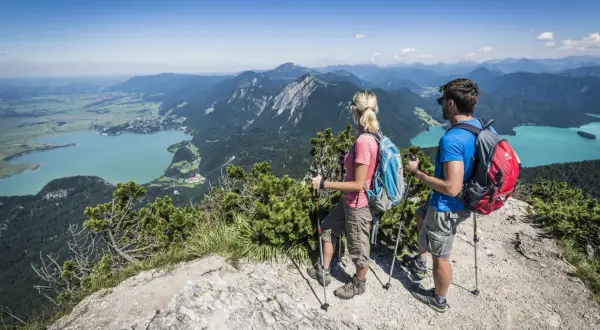 Ein Paar genießt die Aussicht beim Wandern in Mittenwald. Türkisfarbene Seen und grüne Berge erstrecken sich unter ihnen.