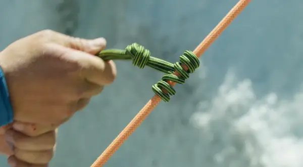 Eine Hand bindet einen HMS Knoten auf ein oranges Seil.